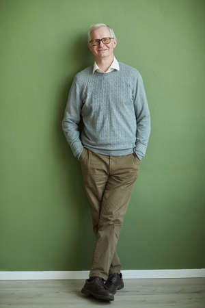 Happy elderly man in eyeglasses, casual brown pants and light blue pullover standing by green wall...