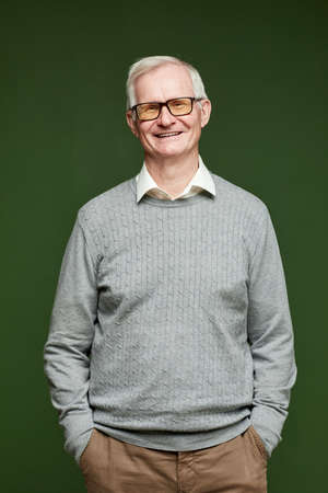 Cheerful elderly man in pants, pullover and eyeglasses standing on dark green background in front of...