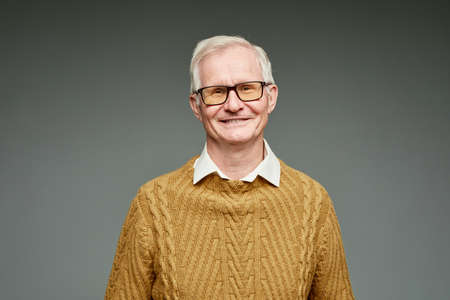 Cheerful aged grey haired man wearing brown knitted sweater and eyeglasses standing against grey...