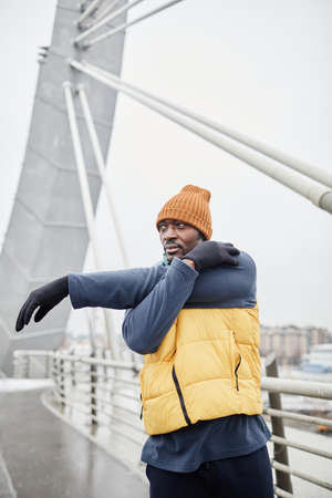 Active young man in sleeveless jacket, cap, gloves and tracksuit stretching arm in front of himself...
