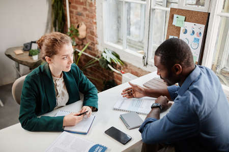 Two young confident intercultural colleagues with business documents discussing working plan or data at working meetingの写真素材