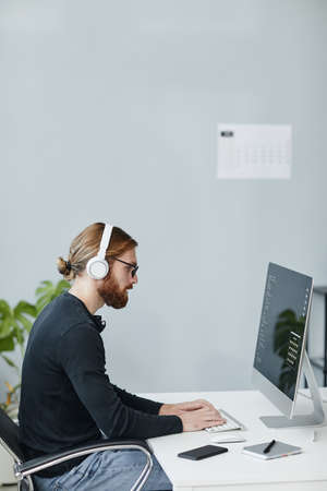 Contemporary young diversity programmer in casual clothes and headphones working in front of...