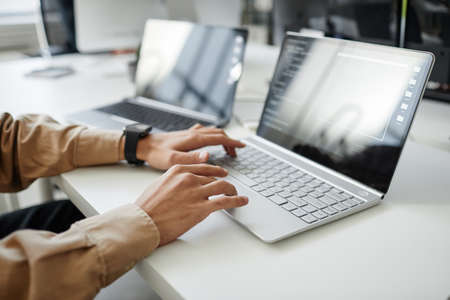Hands of young contemporary program developer typing on laptop keyboard while sitting by desk in...