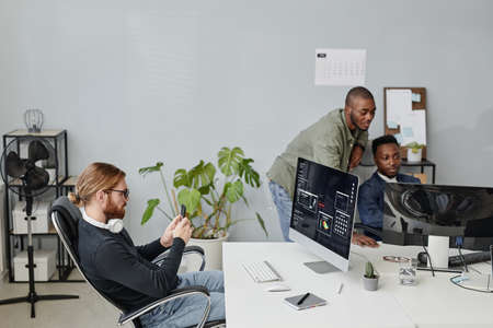 Young businessman with smartphone sitting by desk in front of computer screen against two African American coworkersの写真素材