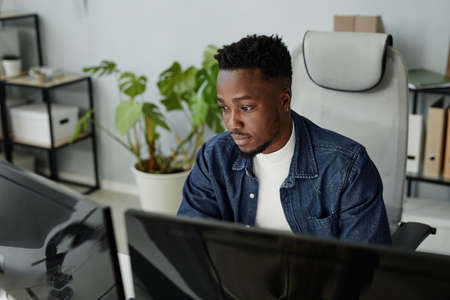 Young blackman in casual clothes sitting in front of computer monitors and looking at data on screens during work in openspace bureauの写真素材