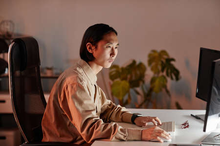 Contemporary young Asian businessman sitting in front of computer monitors while working late in openspace officeの写真素材