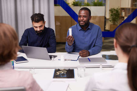 Young confident African-American pharmacologist showing colleagues plastic bottle with pills while presenting new medicamentの写真素材