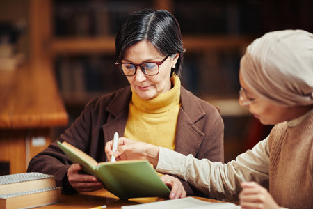 Warm toned portrait of two female adult students reading books while preparing for exam in college libraryの写真素材
