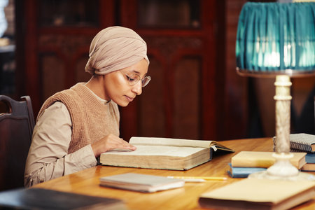 Side view portrait of young Muslim woman reading book in classic college library, copy space
