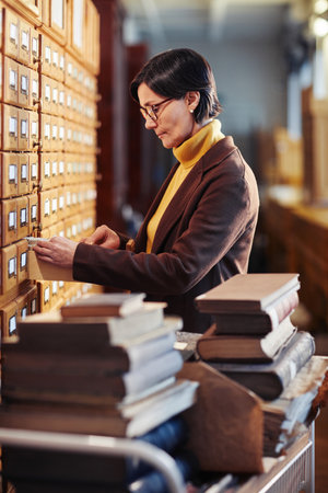Vertical portrait of adult woman searching for book in library catalogue cabinet, copy spaceの写真素材