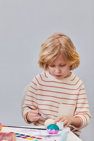 Vertical portrait of cute blonde boy painting Easter eggs while enjoying art and craft class in school against white backgroundの写真素材