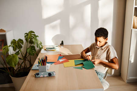 Minimal portrait of young black girl crafting with colored paper at desk lit by sunlight, copy spaceの写真素材