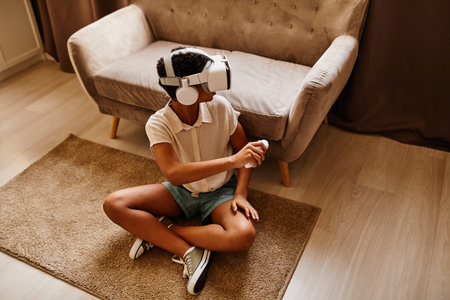 Top view portrait of African American girl playing VR game while sitting on floor at home, copy...