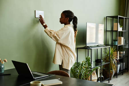 Side view portrait of young African American woman using smart home control panel in minimal modern...