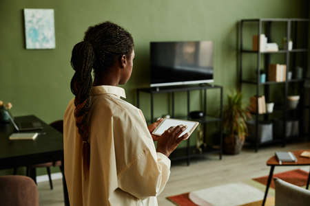 Side view portrait of black young woman holding tablet with blank screen while operating smart home...