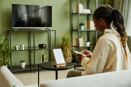 Side view portrait of young black woman connecting phone to smart home system with blank screen,...