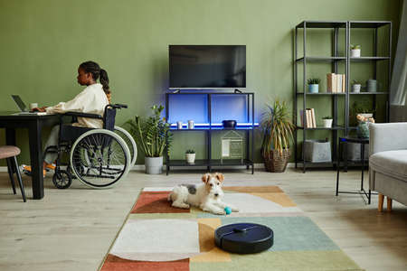 Wide angle view at young African American woman using wheelchair while working at modern home office with smart accessible devices and pet dogの写真素材
