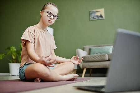 Full length portrait of teenage girl with down syndrome watching online lesson on yoga at home, copy spaceの写真素材