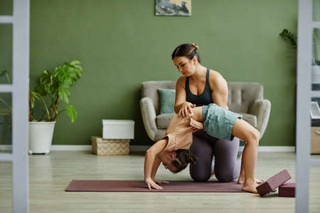 Full length portrait of young female instructor assisting girl with down syndrome exercising in fitness studio and stretching in wheel pose, copy spaceの写真素材