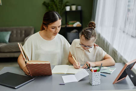 Front view portrait of young female tutor or mother helping girl with down syndrome studying at home...