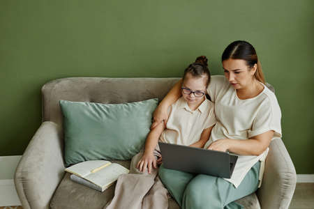 Minimal portrait of mother and daughter with down syndrome relaxing on couch together in cozy home,...