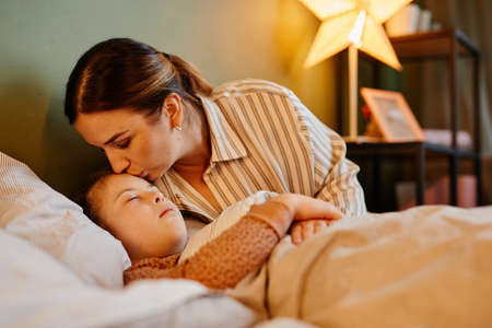 Portrait of caring mother kissing child with down syndrome at bedtime lit by cozy lamp light, copy...