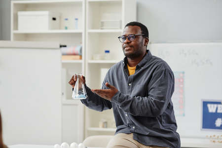 Portrait of African American science teacher explaining chemistry in school class and holding glass...