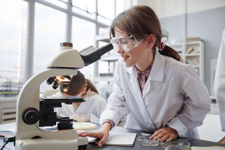 Side view portrait of smiling young girl looking into microscope while enjoying experiments in school chemistry labの写真素材