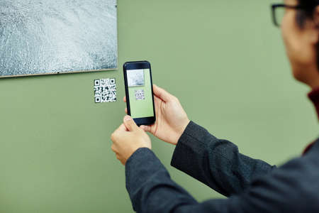 Unrecognizable young man spending time in modern art gallery scanning QR code using his smartphone to get more information about photo on wallの写真素材