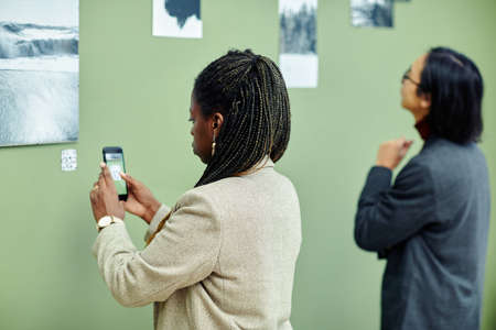 Young African American woman attending exhibition in modern art gallery scanning QR code using smartphone to find more information about photo on wallの写真素材