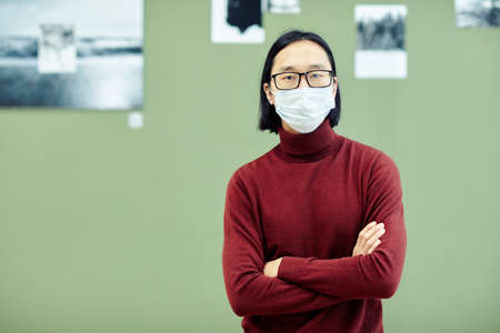 Young Asian man wearing eyeglasses and protective mask on face standing with arms crossed in modern art gallery looking at cameraの写真素材