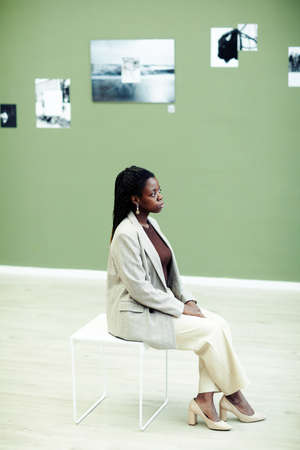 Vertical high angle view of elegant young Black woman sitting on stool in contemporary art gallery...