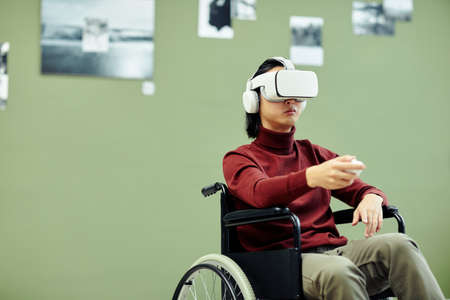 Horizontal medium shot of young Asian man with disability wearing VR headset visiting exhibition in modern art gallery using augmented reality technologyの写真素材