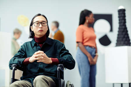 Portrait of handsome young Asian man with disability wearing eyeglasses attending contemporary art exhibition sitting in wheelchair looking at cameraの写真素材
