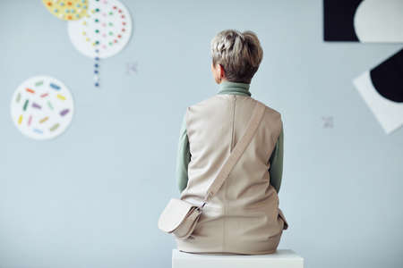 Rear view of unrecognizable mature woman sitting on stool in modern art gallery looking at artworksの写真素材