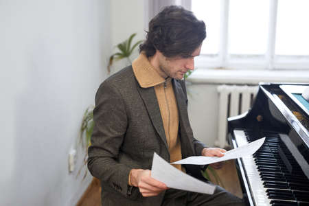 Young composer in jacket sitting opposite the grand piano and examining the sheet music before...