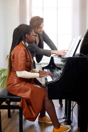 Young man pointing at sheet music and teaching woman to play the piano during lesson at classの写真素材