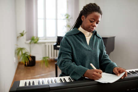African young woman making notes on paper while sitting at synthesizer, she composing the music at...