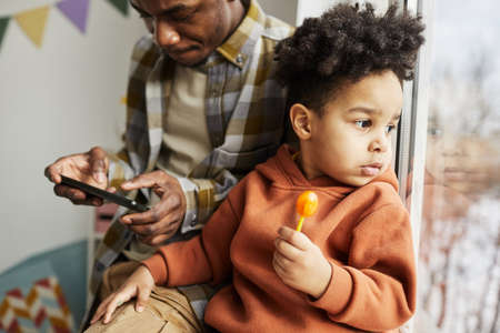 Portrait of cute little boy holding lollipop and looking at window with father in background, copy...