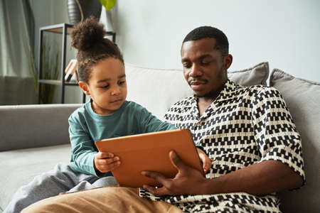 Portrait of young black father and little boy using digital tablet together while relaxing on couch...