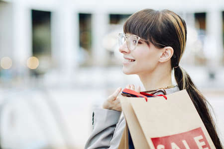 Clean side view portrait of smiling young woman holding shopping bags in mall and wearing glasses,...