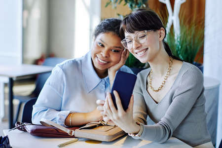 Candid portrait of two young girls looking at smartphone screen at cafe table and enjoying day out