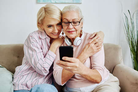 Portrait of modern senior woman using smartphone device at home with smiling young daughter embracing herの写真素材