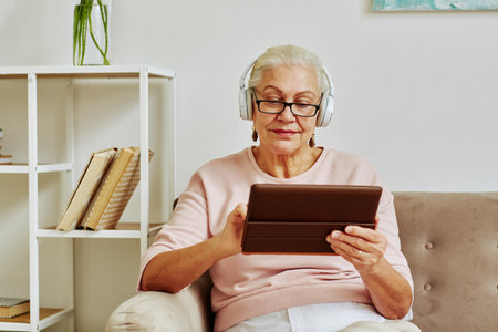 Portrait of modern senior woman using digital tablet with headphones while sitting on sofa in minimal home interiorの写真素材