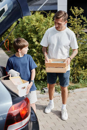 Vertical portrait of father and son carrying boxes to car while moving into new house lit by sunlight outdoorsの写真素材