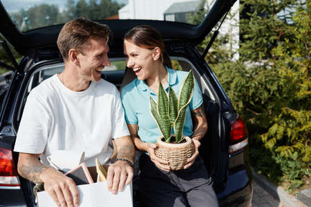 Portrait of adult couple sitting in car trunk with boxes and looking at each other while moving to new houseの写真素材