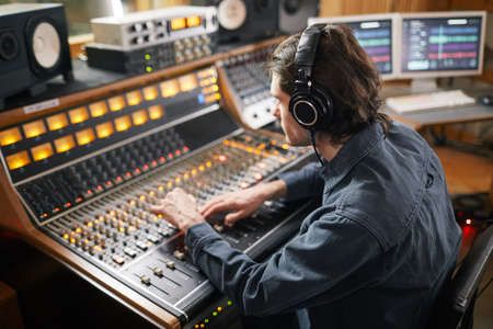 Portrait of young man wearing headphones and operating buttons and toggles at digital audio workstation in recording studio, music production conceptの写真素材