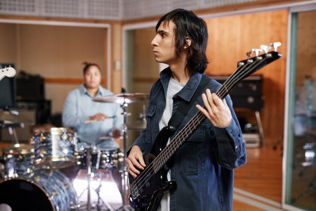 Side view portrait of young man playing bass guitar while recording new tracks with band in...