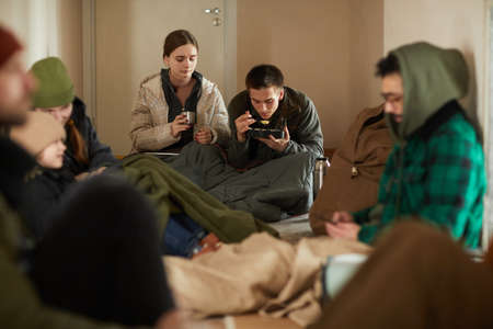 Portrait of Caucasian young couple eating simple food while hiding in refugee shelter with group of...