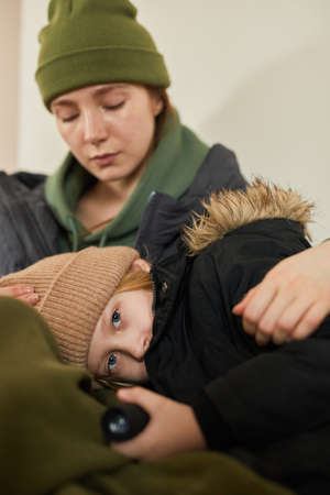 Vertical portrait of blonde Caucasian child lying in mothers lap at refugee shelter and looking at...